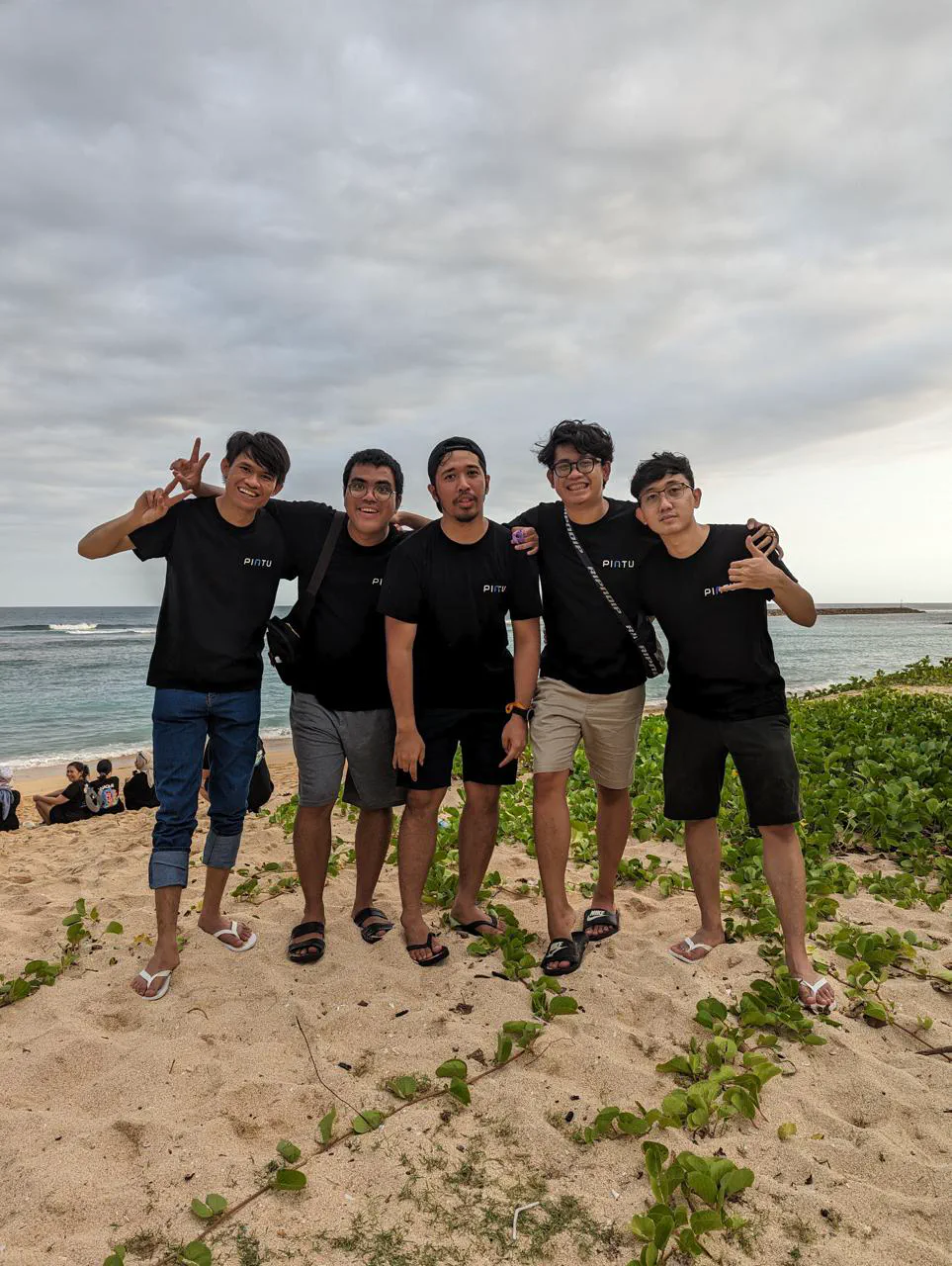 Five people in black Pintu t-shirts with their arms around each other on a beach.