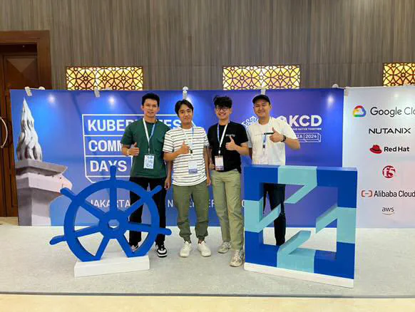 Four people with conference lanyards giving a thumbs up in front of the Kubernetes Community Days Indonesia 2024 backdrop.