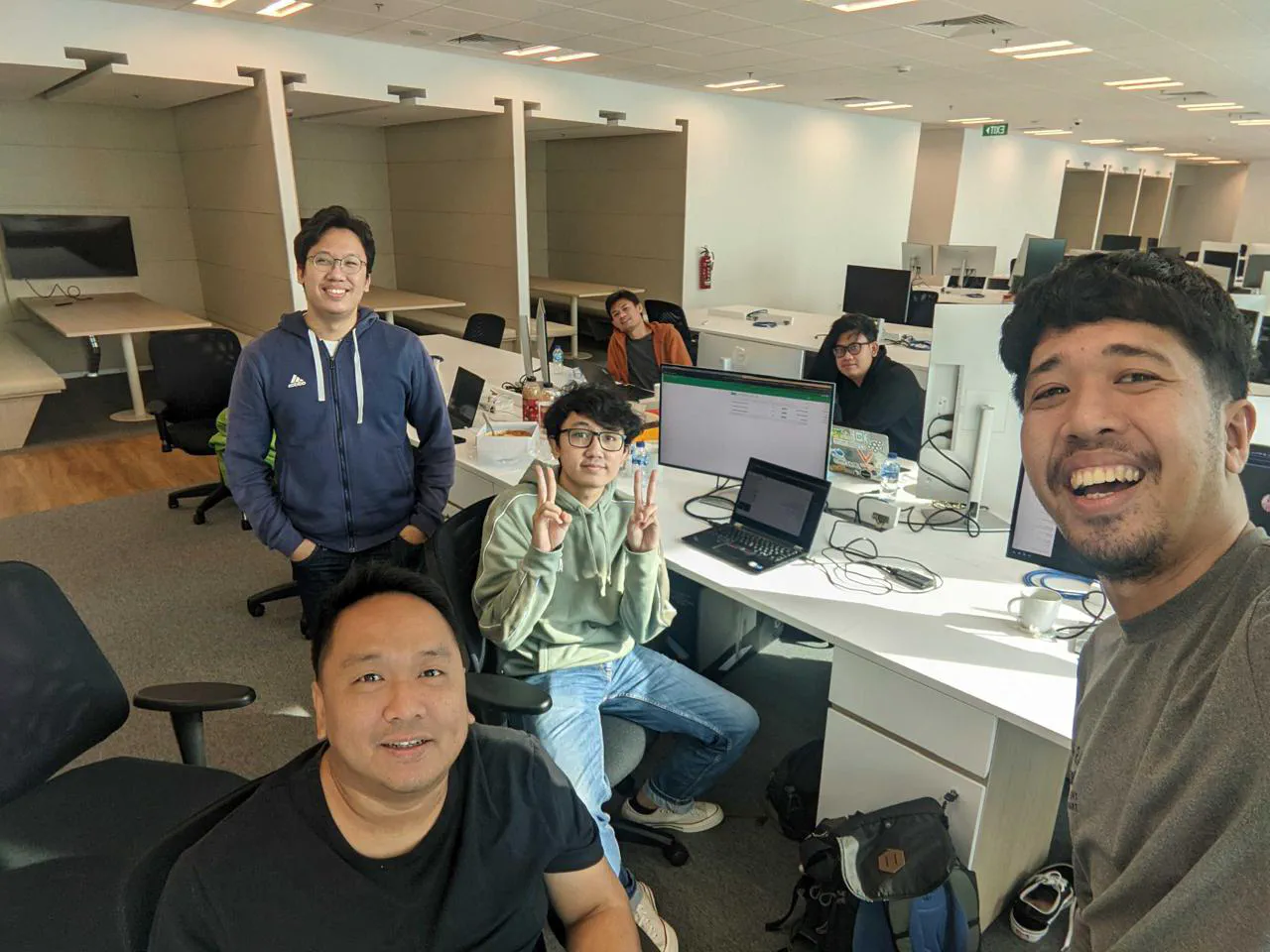 Selfie of six people at their desks in a mostly empty open-plan office, with laptops and monitors.