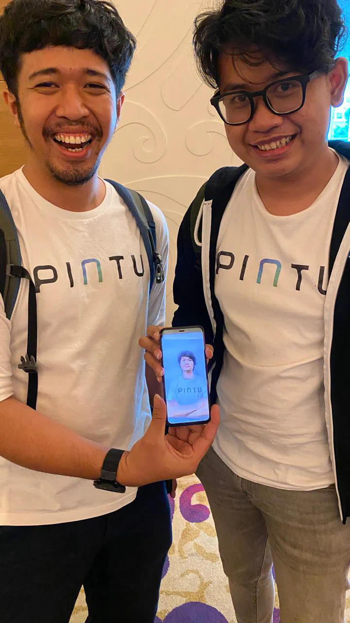 Two people in white Pintu t-shirts smiling. One holds up a phone showing a photo of someone else in a Pintu t-shirt.