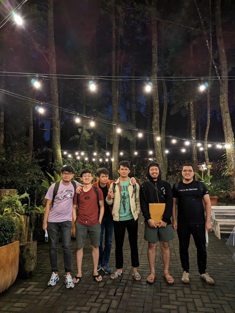 Six people standing at night under string lights among tall pine trees.