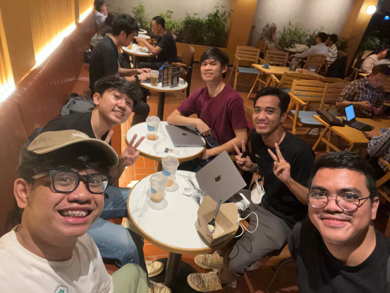 Selfie of five people at a café at night, with laptops and iced drinks on small round tables.
